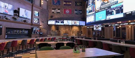 A sports bar with multiple screens displaying various sports events, a bar area with a selection of bottles, and seating arrangements for patrons.
