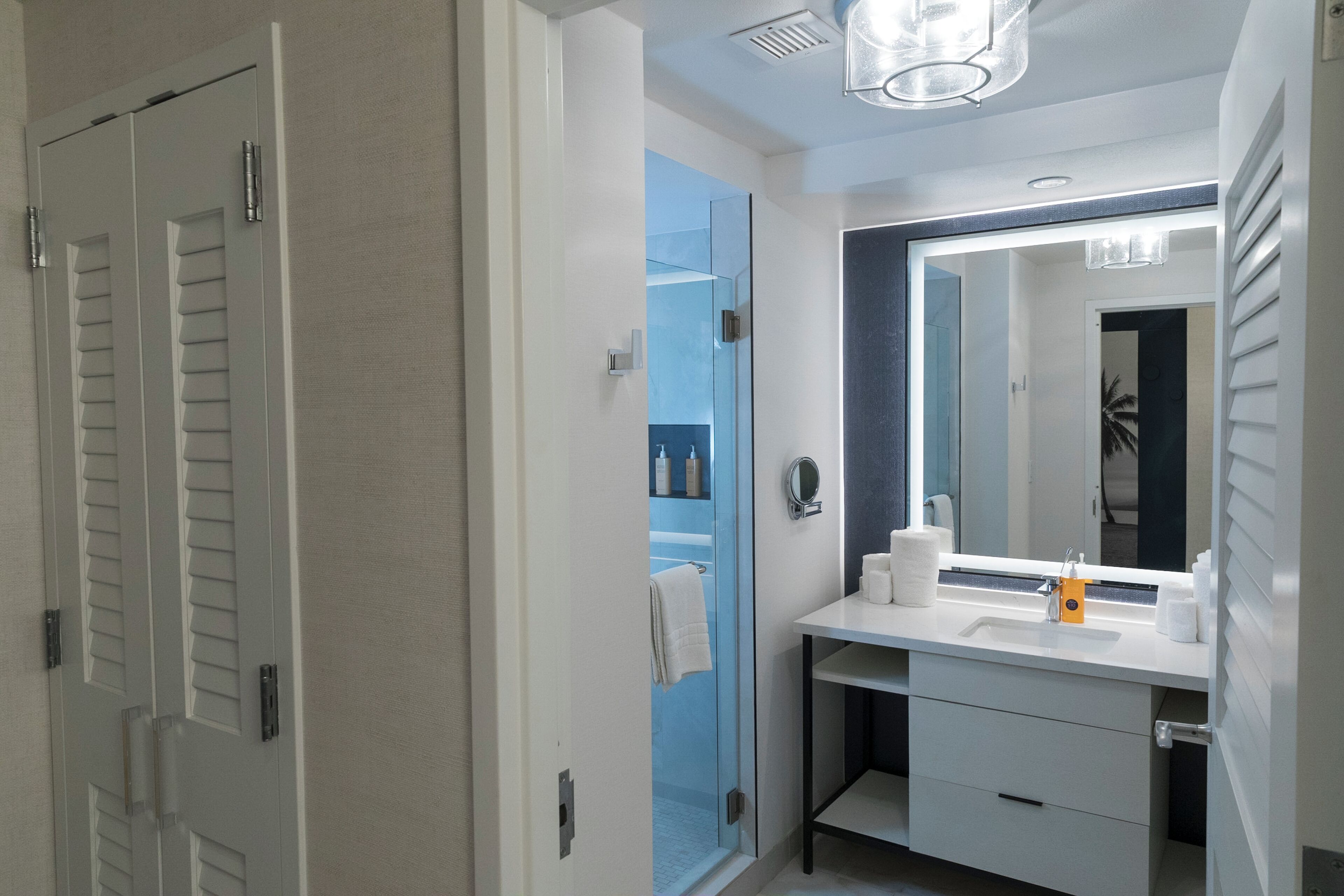 Un baño moderno con cabina de ducha de vidrio, un tocador blanco con un espejo grande, y una toalla colgada en la puerta.