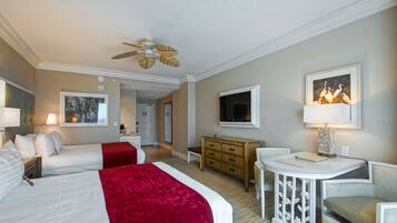A hotel room with two beds, a desk, a chair, a TV, a ceiling fan, and artwork on the walls.