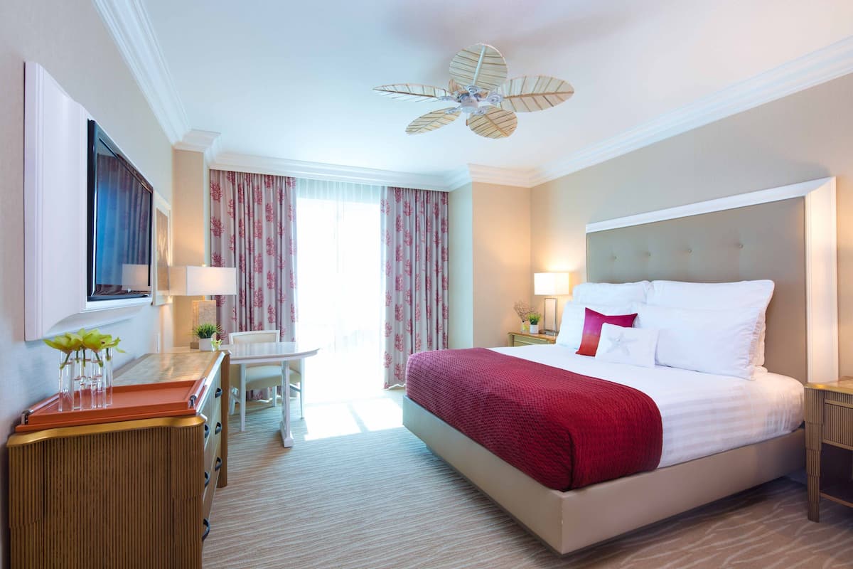 A hotel room with a large bed, a desk with a chair, a ceiling fan, and a window with curtains.