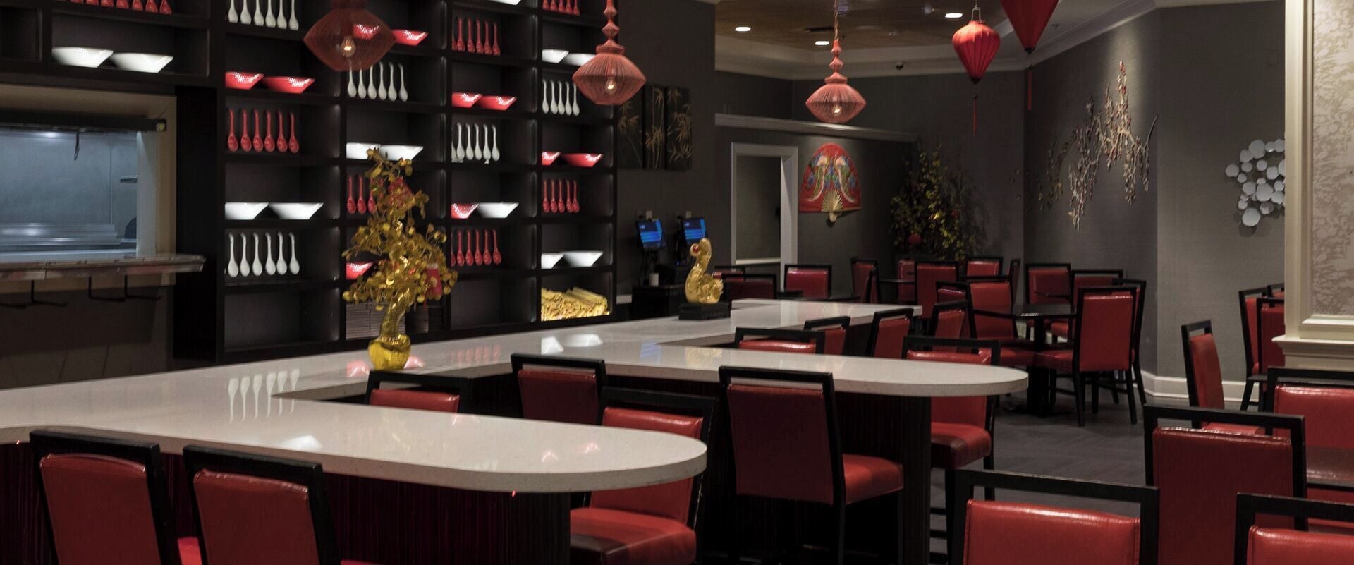 Een restaurant met een centraal gelegen bar, rode bekleding op de stoelen en decoratieve elementen zoals een gouden beeld en rode lantaarns.