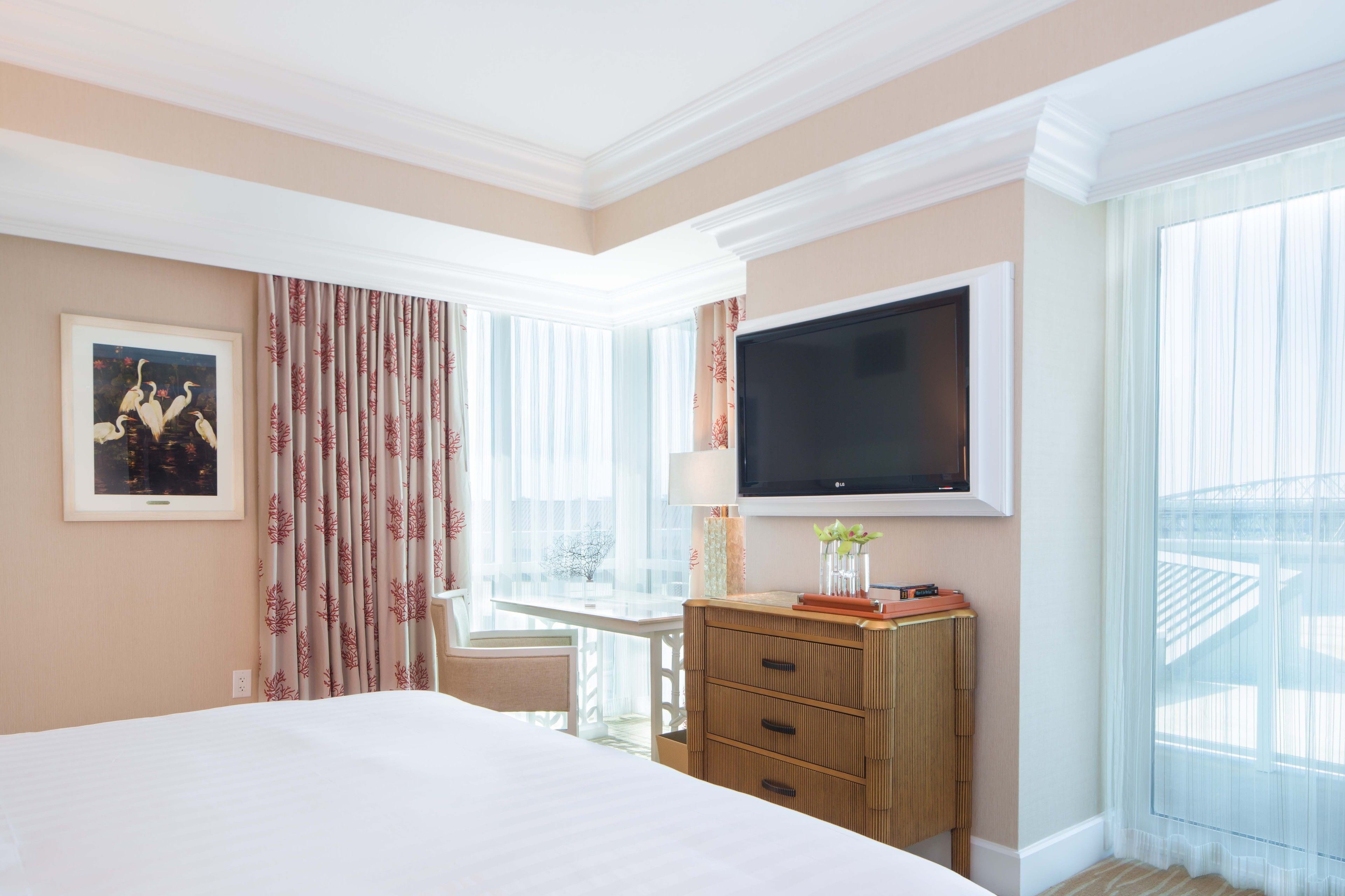 A hotel room with a large bed, a television mounted on the wall, a wooden dresser, and a window with sheer curtains.