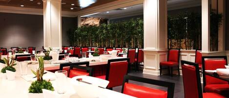 A restaurant with red chairs and white tables, large pillars, and potted plants.