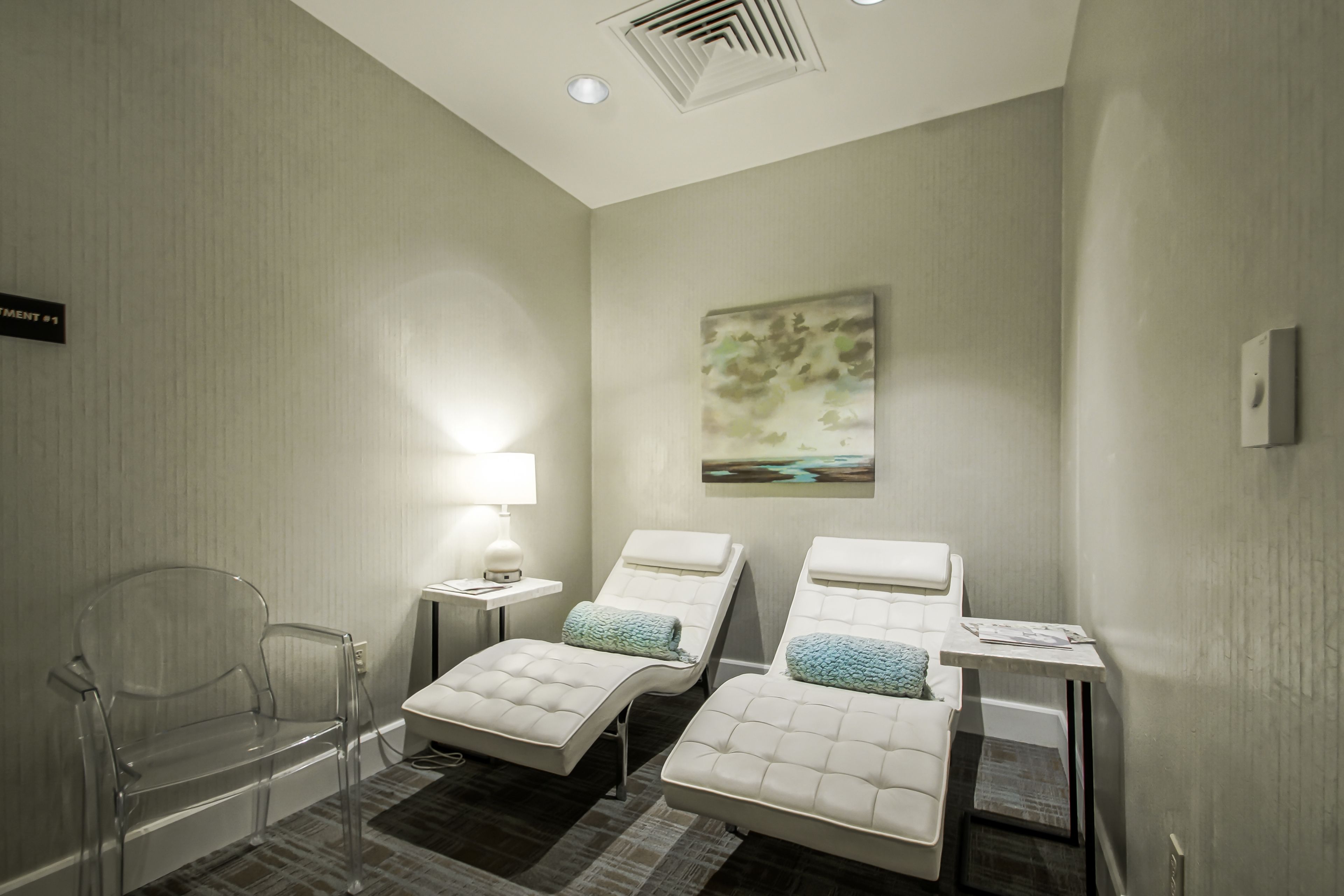 Une pièce comprenant deux fauteuils inclinables, une petite table avec une lampe, un tableau au mur et un panneau indiquant le numéro de la pièce.
