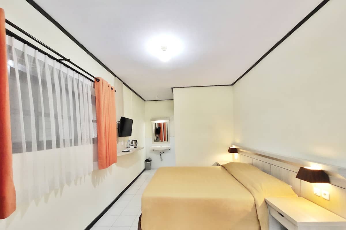 Quarto de hotel com cama, mesa de cabeceira, luminária, televisão e banheiro com pia e espelho.