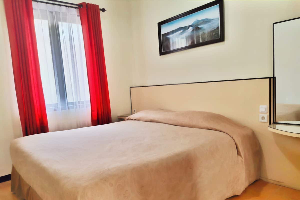 Cama bem arrumada com colcha bege em quarto de hotel, parede lisa, quadro emoldurado e janela com cortinas vermelhas.