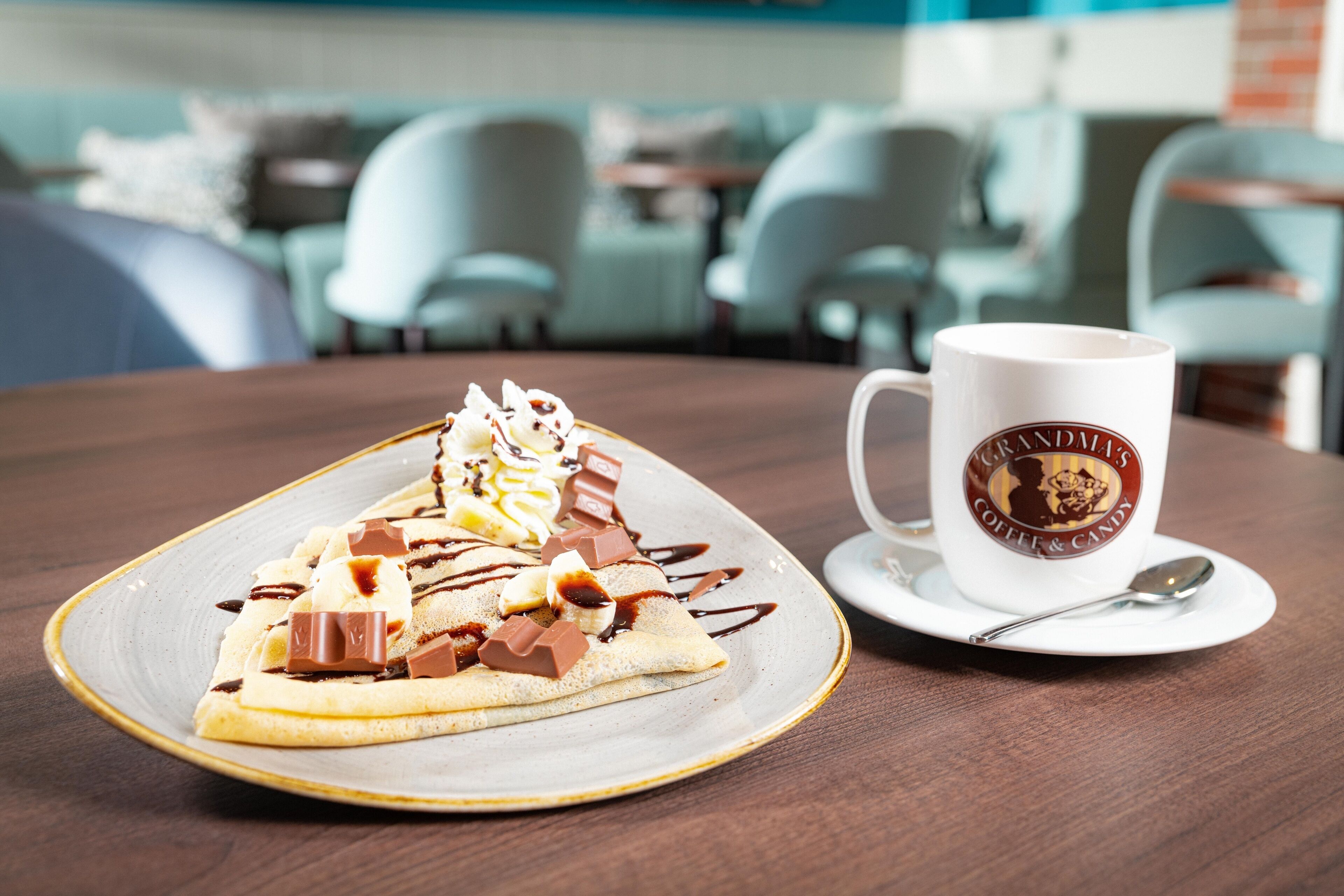 Un plato de postre con un crepe cubierto de crema batida, trozos de chocolate y salsa de caramelo, acompañado de una taza de café con una cuchara sobre una mesa en un café.