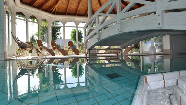 Una piscina interior con paredes de vidrio transparente, techo de madera y sillas de descanso a lo largo del borde.
