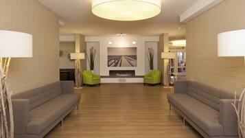 A spacious lobby with a wooden floor, two grey sofas, green chairs, a fireplace, and a large painting on the wall.