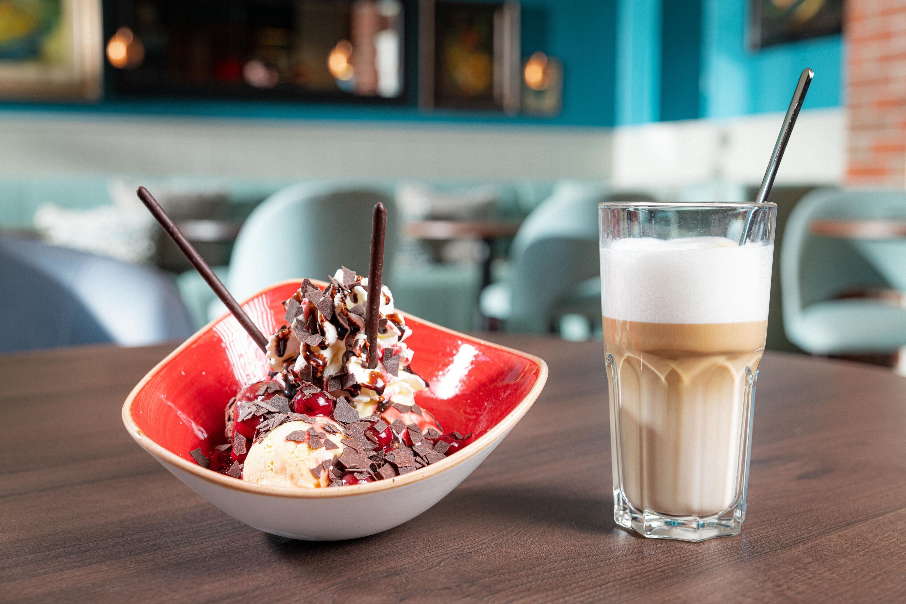 A bowl of dessert with ice cream and chocolate pieces, next to a glass of milkshake on a wooden table.
