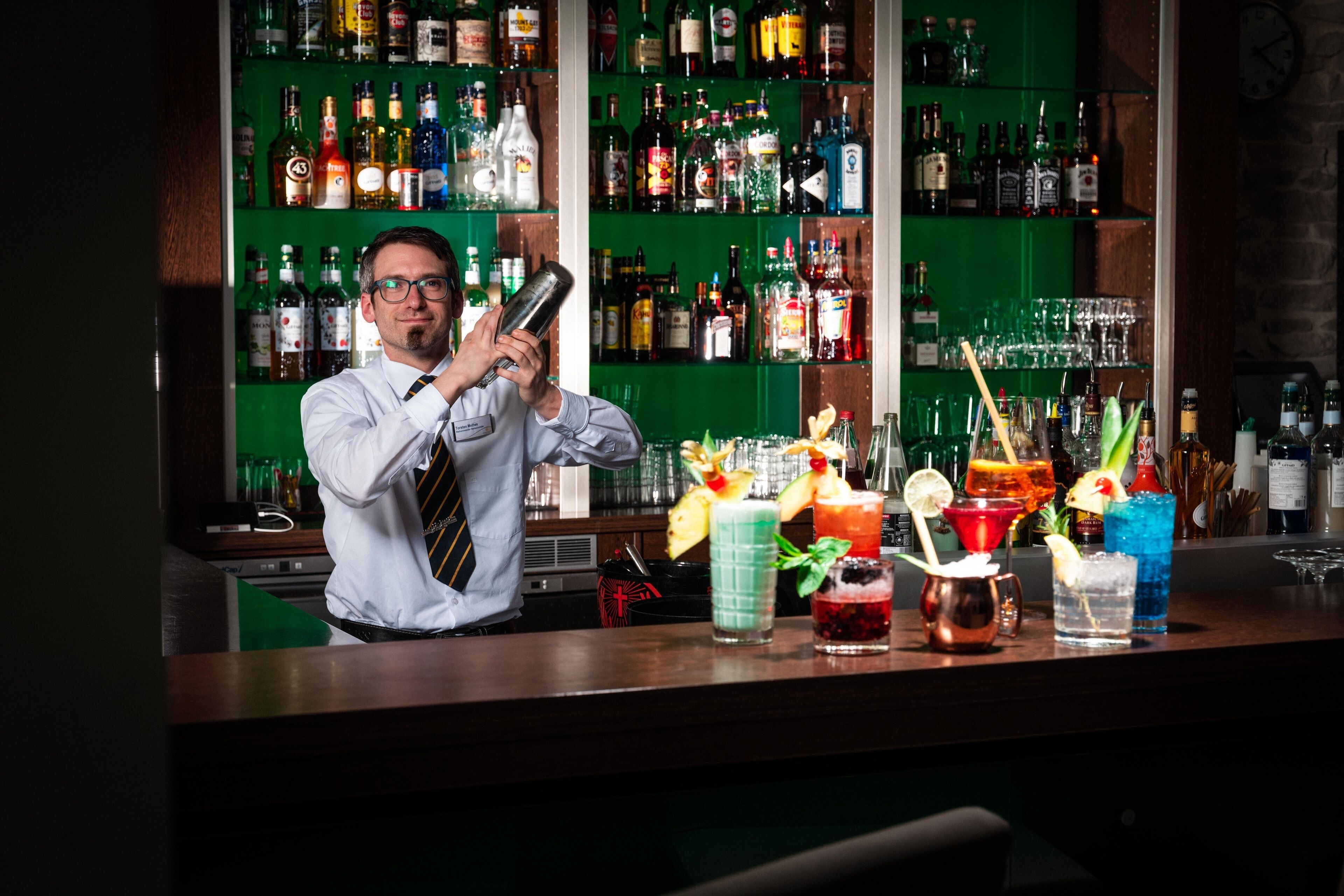 A bartender preparing drinks at a bar with a green backdrop and shelves of liquor bottles.