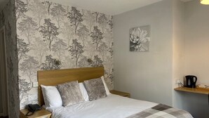 Un lit bien fait, recouvert d’une couverture à carreaux, une table de chevet en bois avec un téléphone, et un mur orné d’un papier peint à motifs floraux.