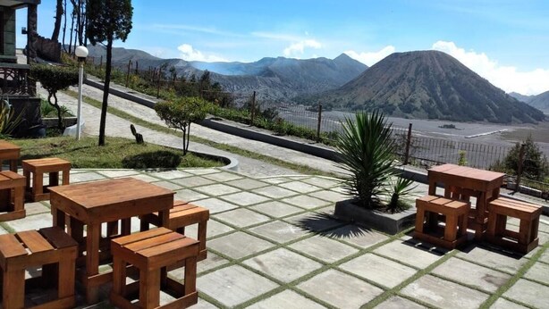 Un área de estar al aire libre con mesas y sillas de madera, un camino pavimentado y vistas a una cadena montañosa.