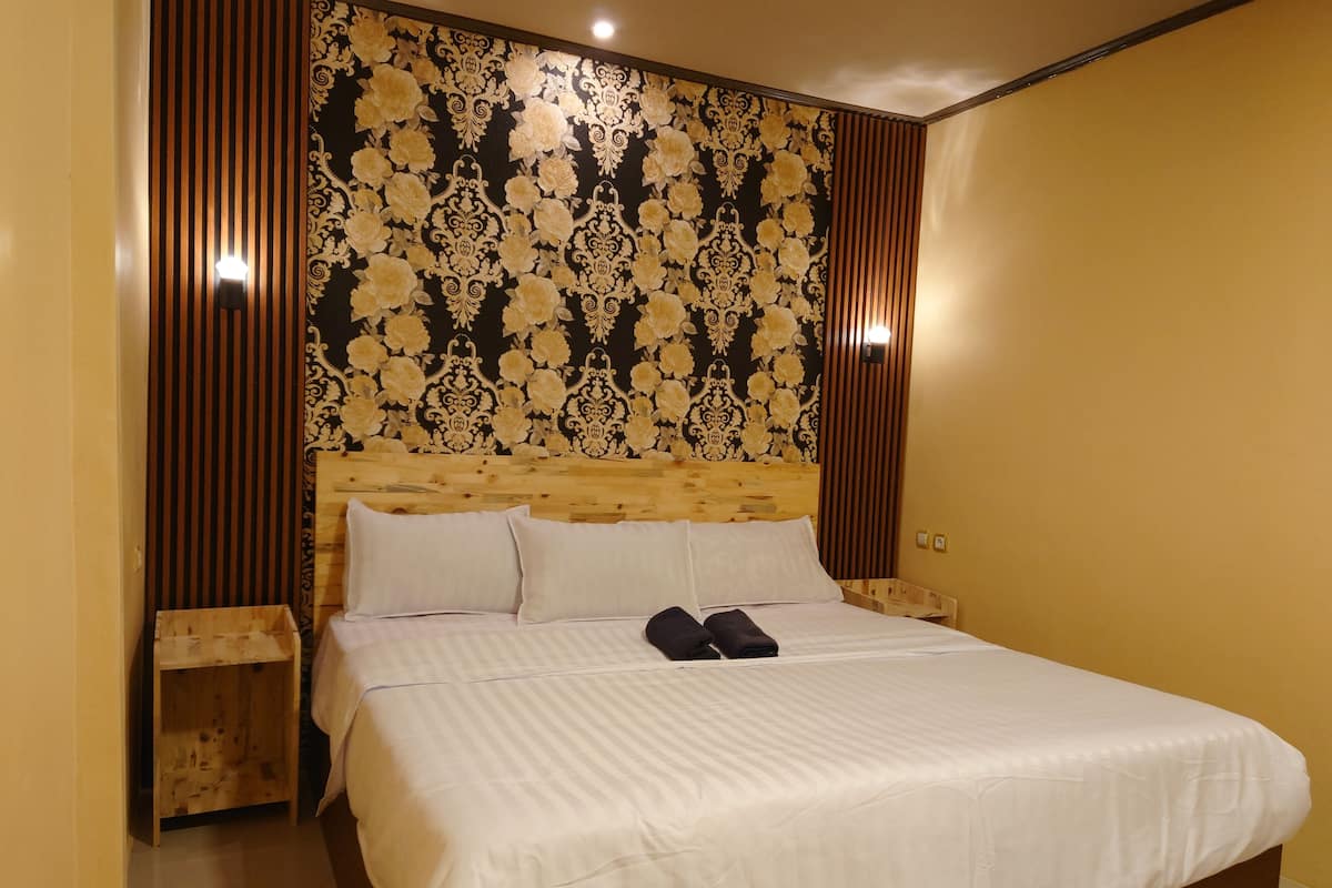 A neatly made bed with white linens and two folded towels, a wooden headboard with a floral pattern, and a small wooden nightstand beside the bed.