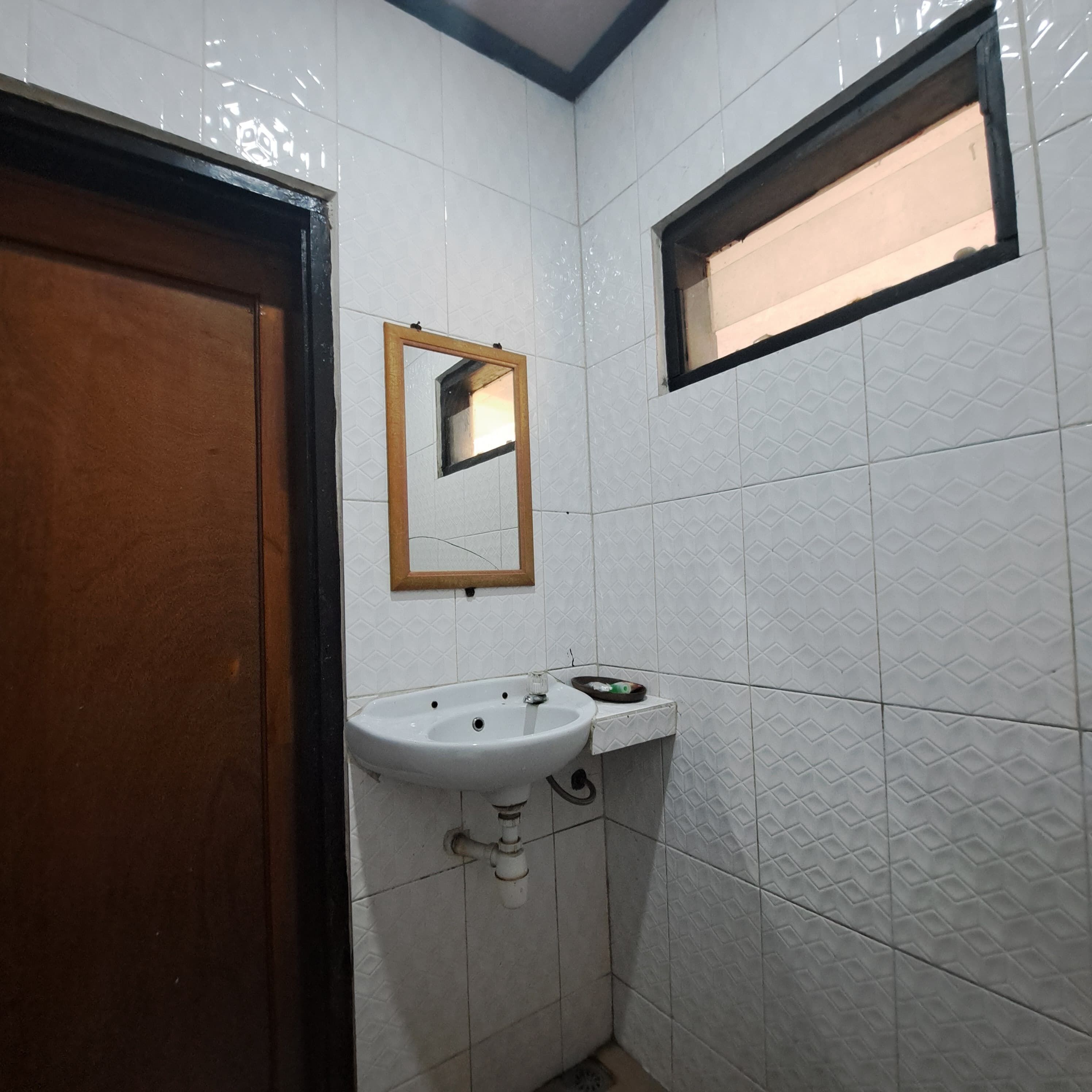 Ein kleines Badezimmer mit weiß gefliesten Wänden, einer Holztür, einem weißen Waschbecken mit Spiegel darüber und einem Fenster mit schwarzem Rahmen.