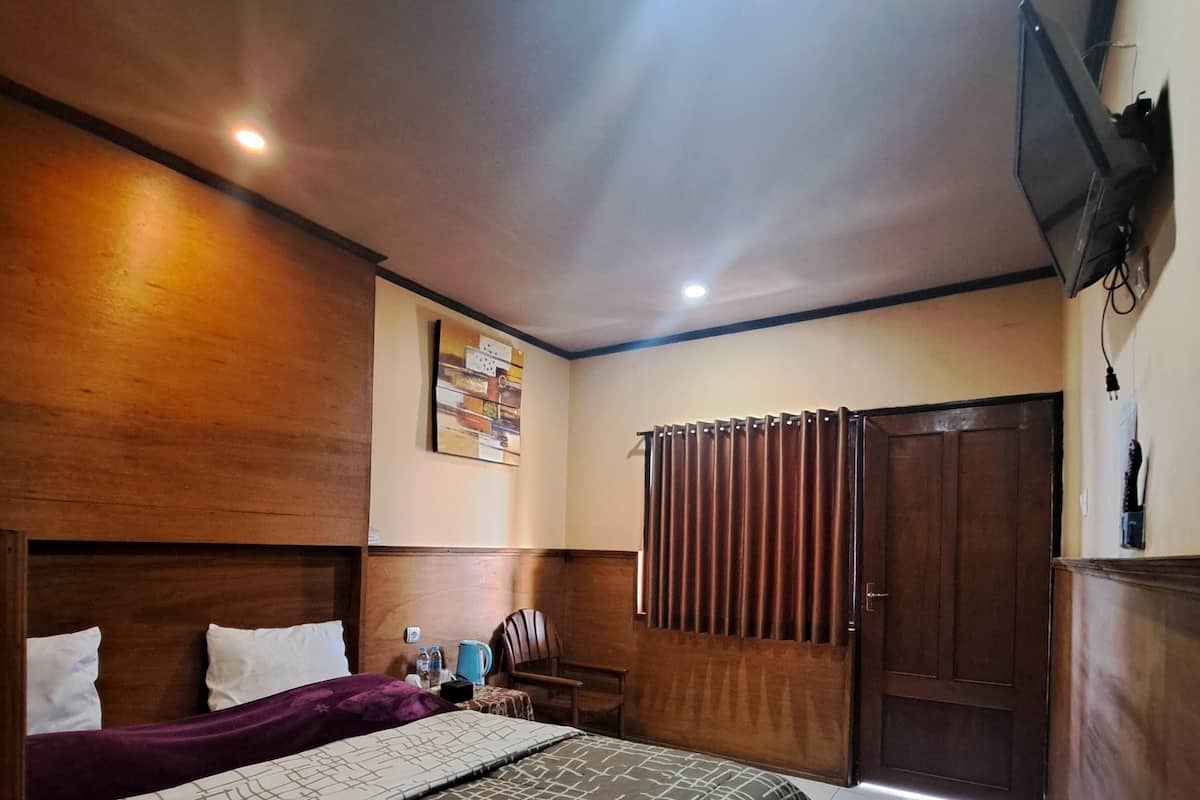 A hotel room with a bed, a wooden headboard, a television mounted on the wall, a door with a curtain, and a small table with a kettle and water bottles.