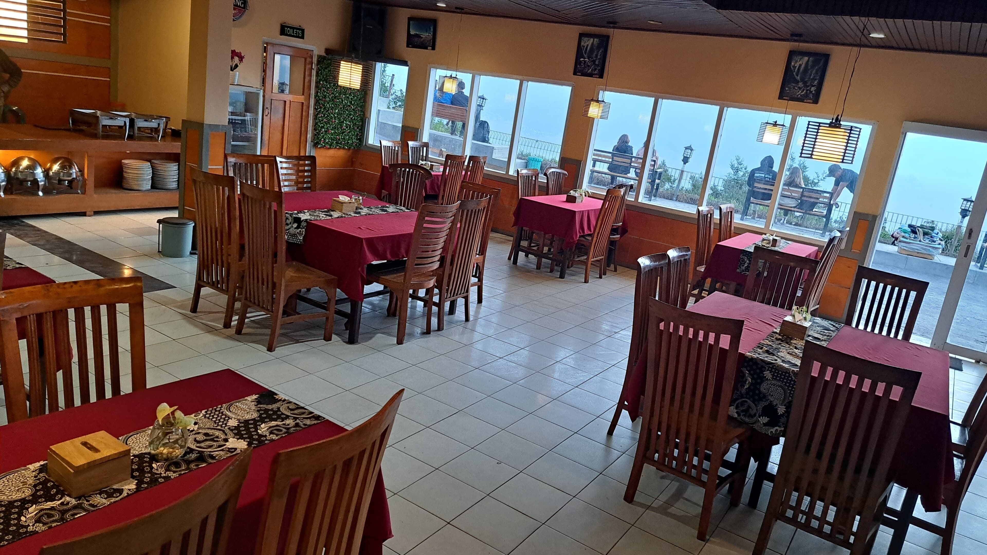 A restaurant with multiple tables and chairs, a visible sign for toilets, and large windows offering a view.