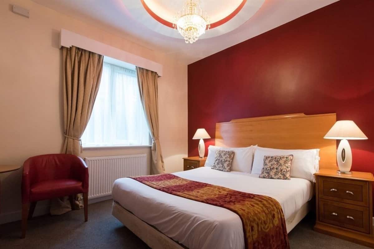 A hotel room with a large bed, two bedside lamps, a red chair, and a window with curtains.