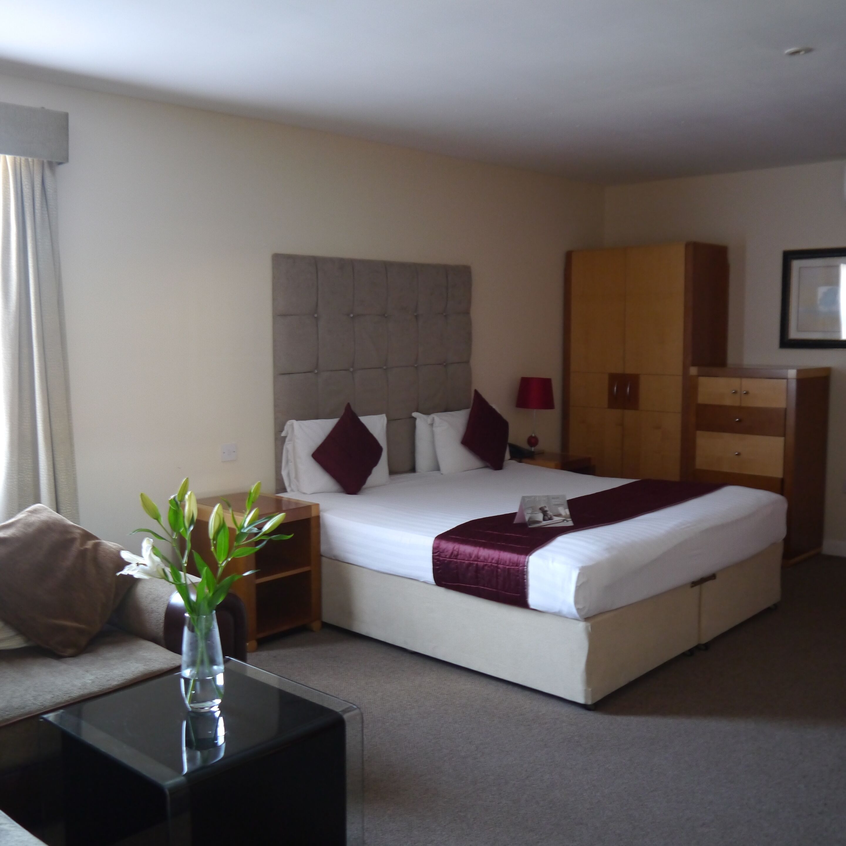 A hotel room with a large bed, a nightstand, a dresser, and a small table with a vase of flowers.