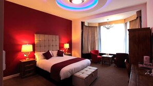 Une chambre d’hôtel avec un grand lit, deux tables de chevet avec des lampes, un mur d’accent rouge, un lustre, un coin repas avec une table et des chaises, et une vue sur l’extérieur.
