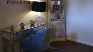 Une pièce comprenant un bureau, une chaise bleue, une lampe, un meuble décoratif contenant des bouteilles et une enseigne murale indiquant « THE PEARL HOTEL ».