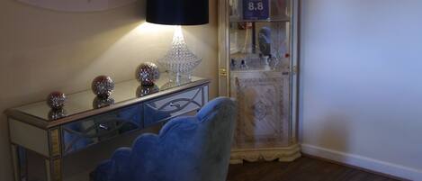 Una habitación con un escritorio, una silla azul, una lámpara, un aparador decorativo con botellas y un cartel en la pared que dice "THE PEARL HOTEL".