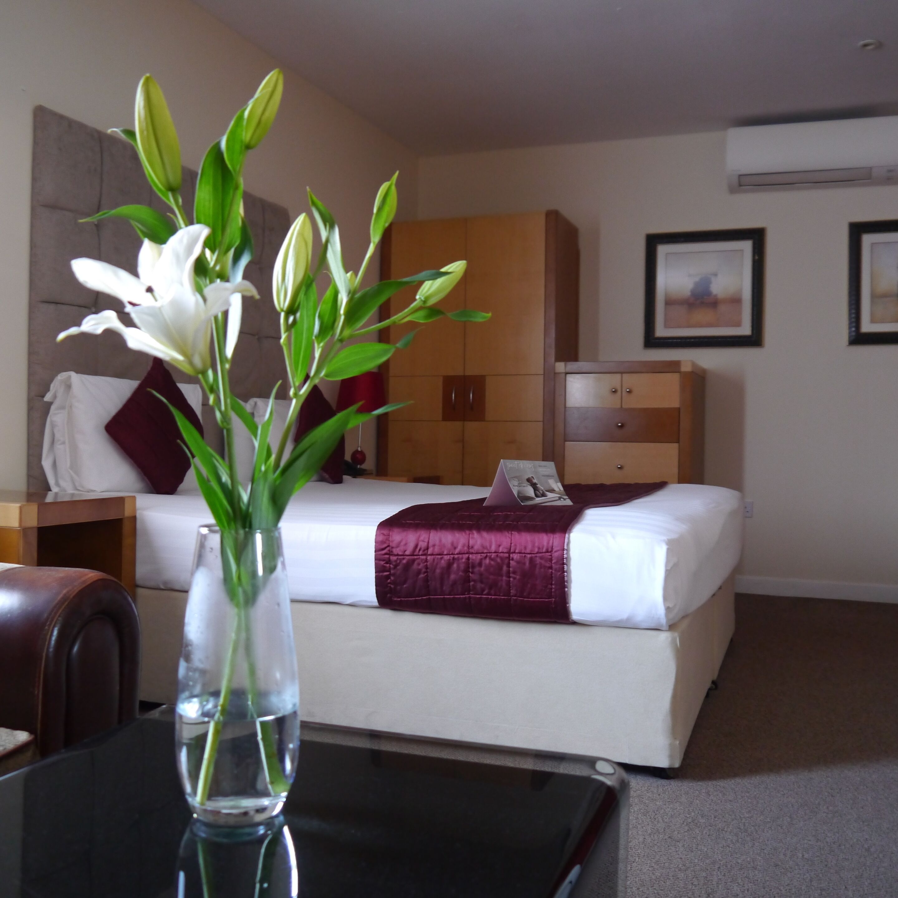 A hotel room with a bed, a nightstand, a desk with a vase of flowers, and a wall-mounted air conditioner.