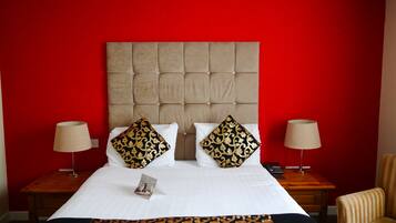 Une chambre d’hôtel avec un mur rouge, une tête de lit capitonnée, deux tables de chevet avec des lampes, une chaise à rayures et un lit avec du linge de lit blanc et des oreillers à motifs.