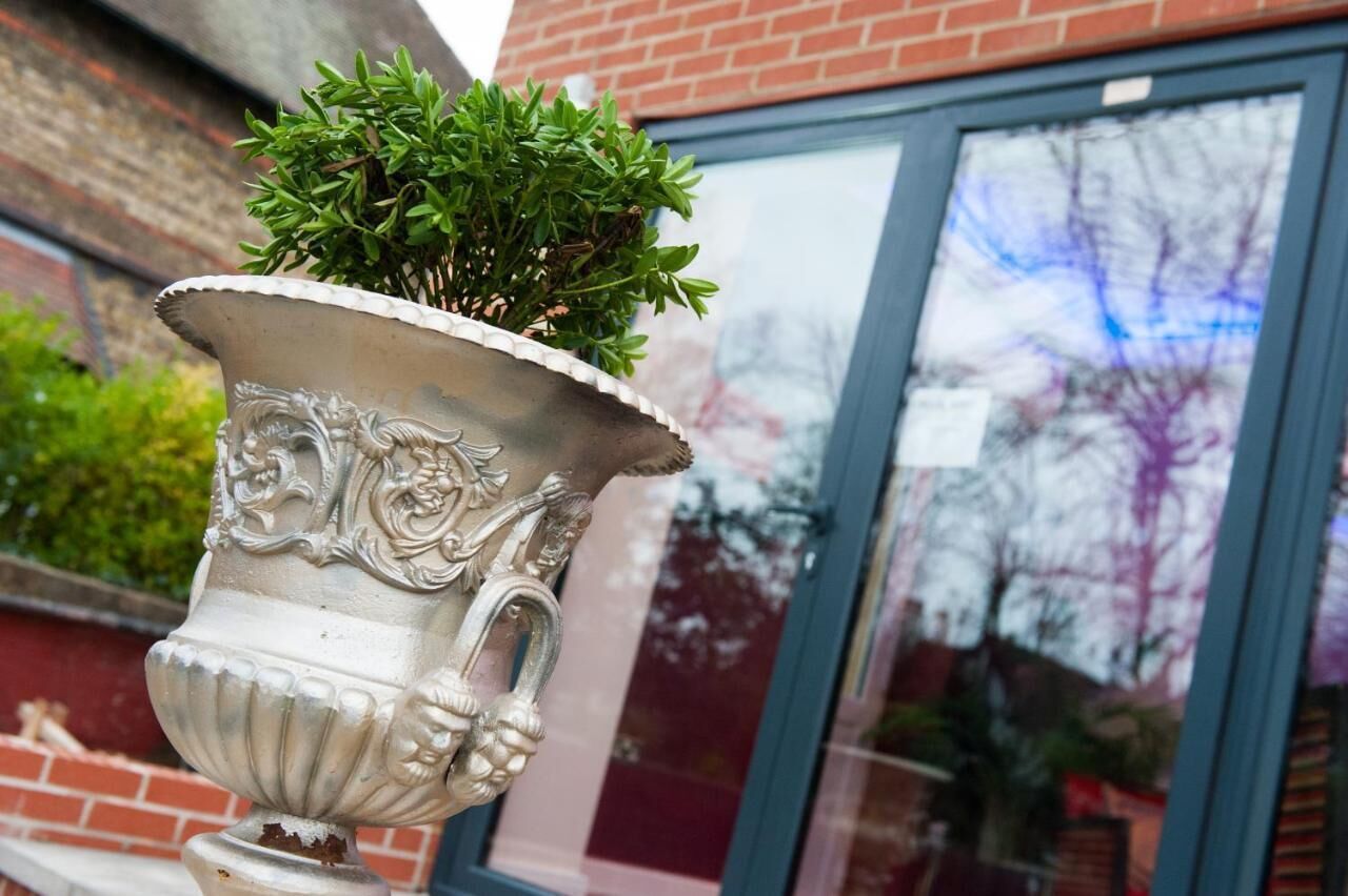 A silver urn with a plant, placed in front of a building with a brick facade and a window with a reflection.
