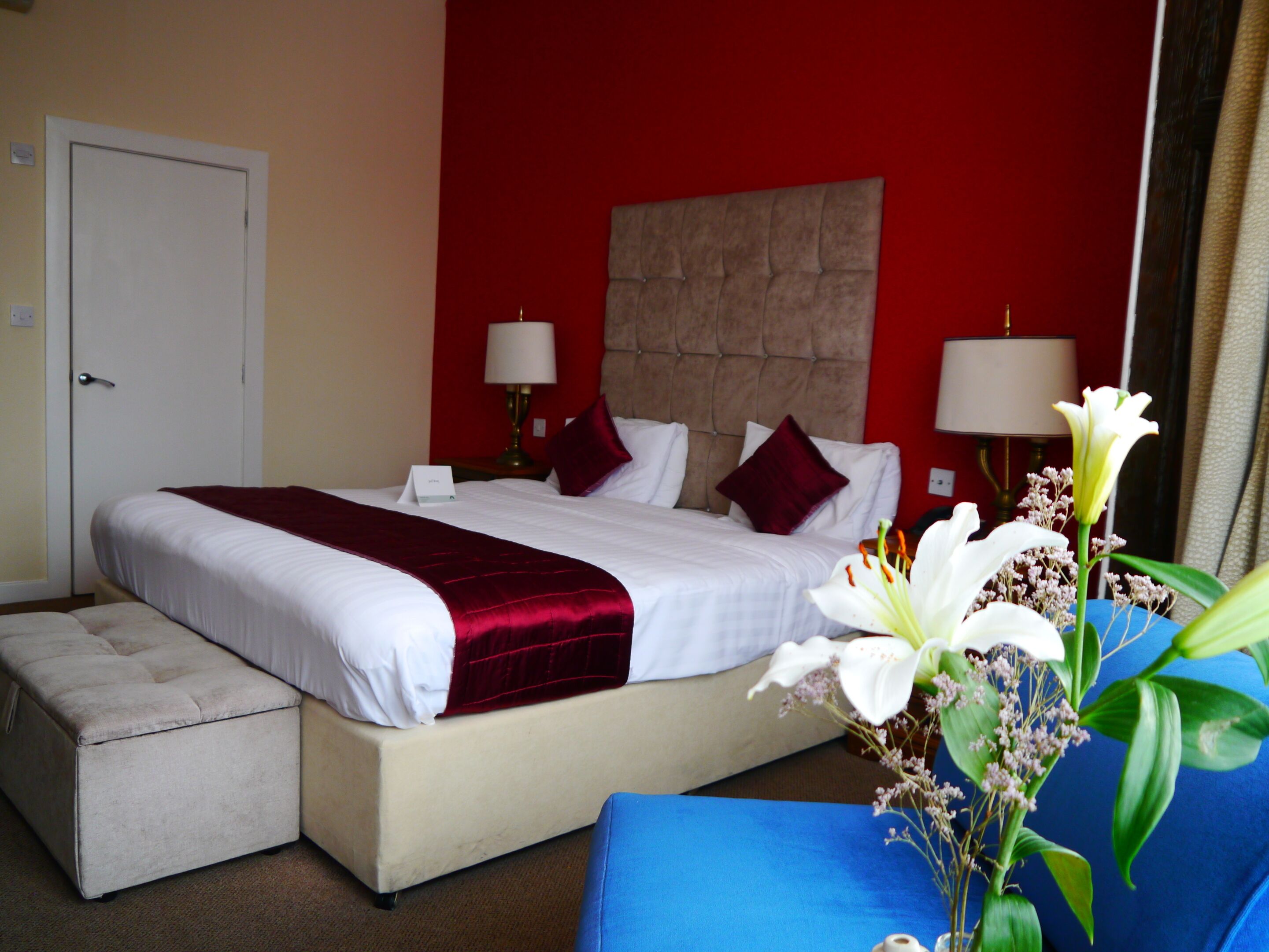 A hotel room with a large bed, a bedside table with lamps, a small bedside table with a lamp, a red accent wall, and a blue chair with flowers.