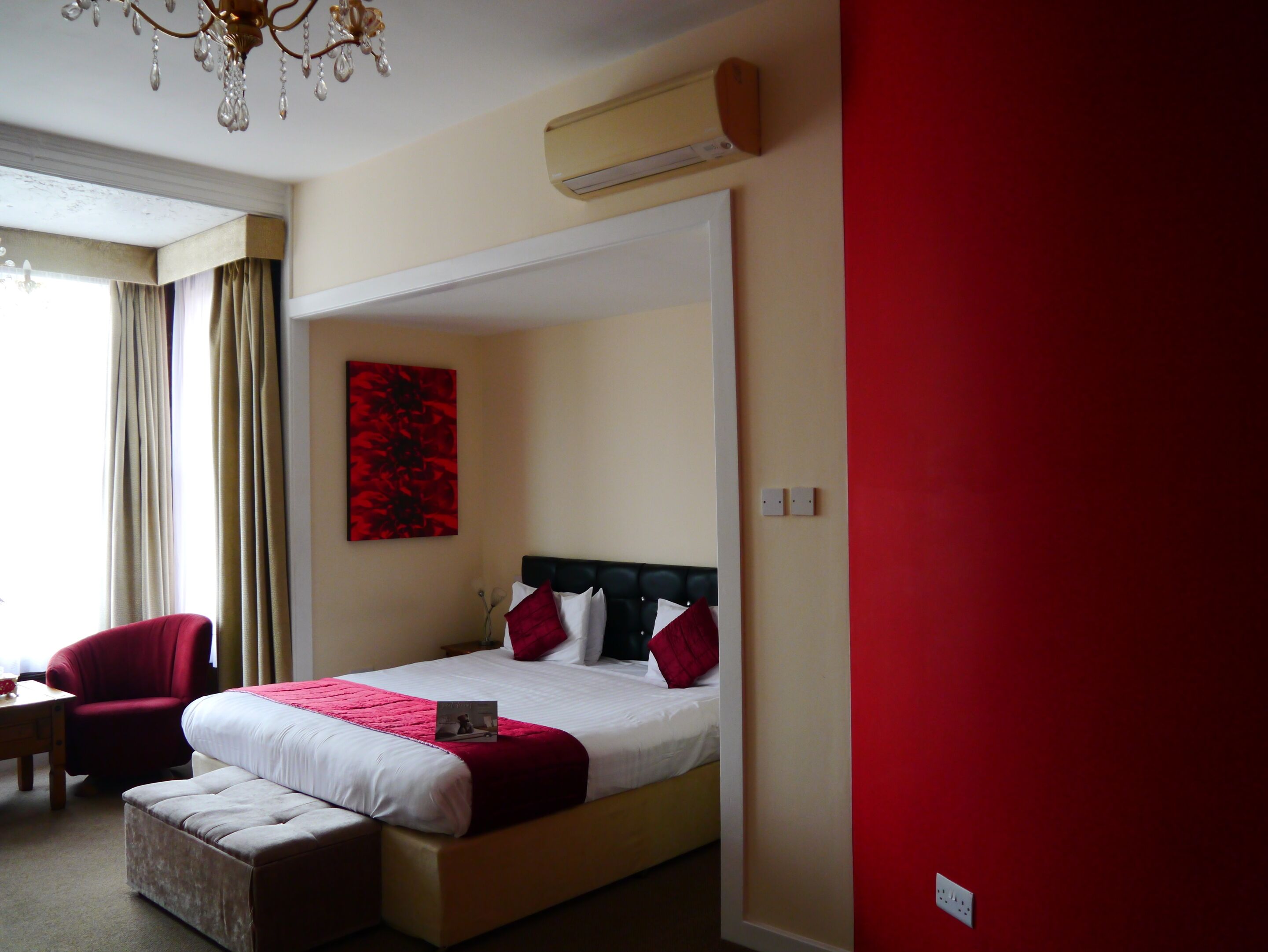 A hotel room with a bed, a red wall, a chandelier, and a window with curtains.
