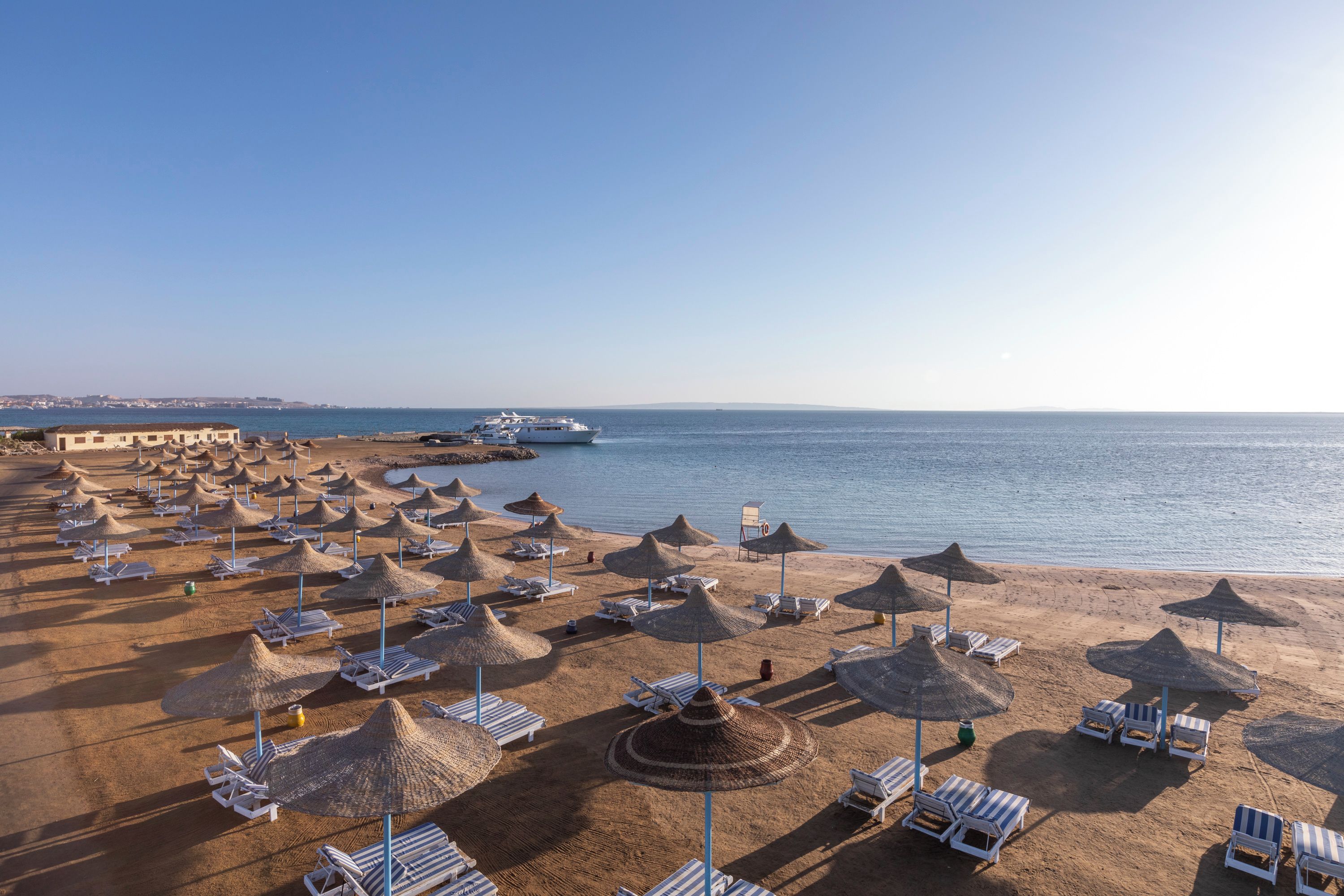 A beach with rows of sun loungers and thatched umbrellas, a yacht docked near the shore, and a clear view of the sea.