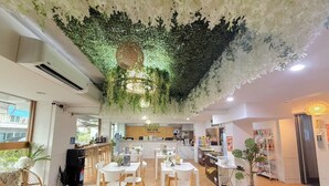 Un restaurante con mesas y sillas blancas, piso de madera y plantas colgantes en el techo.