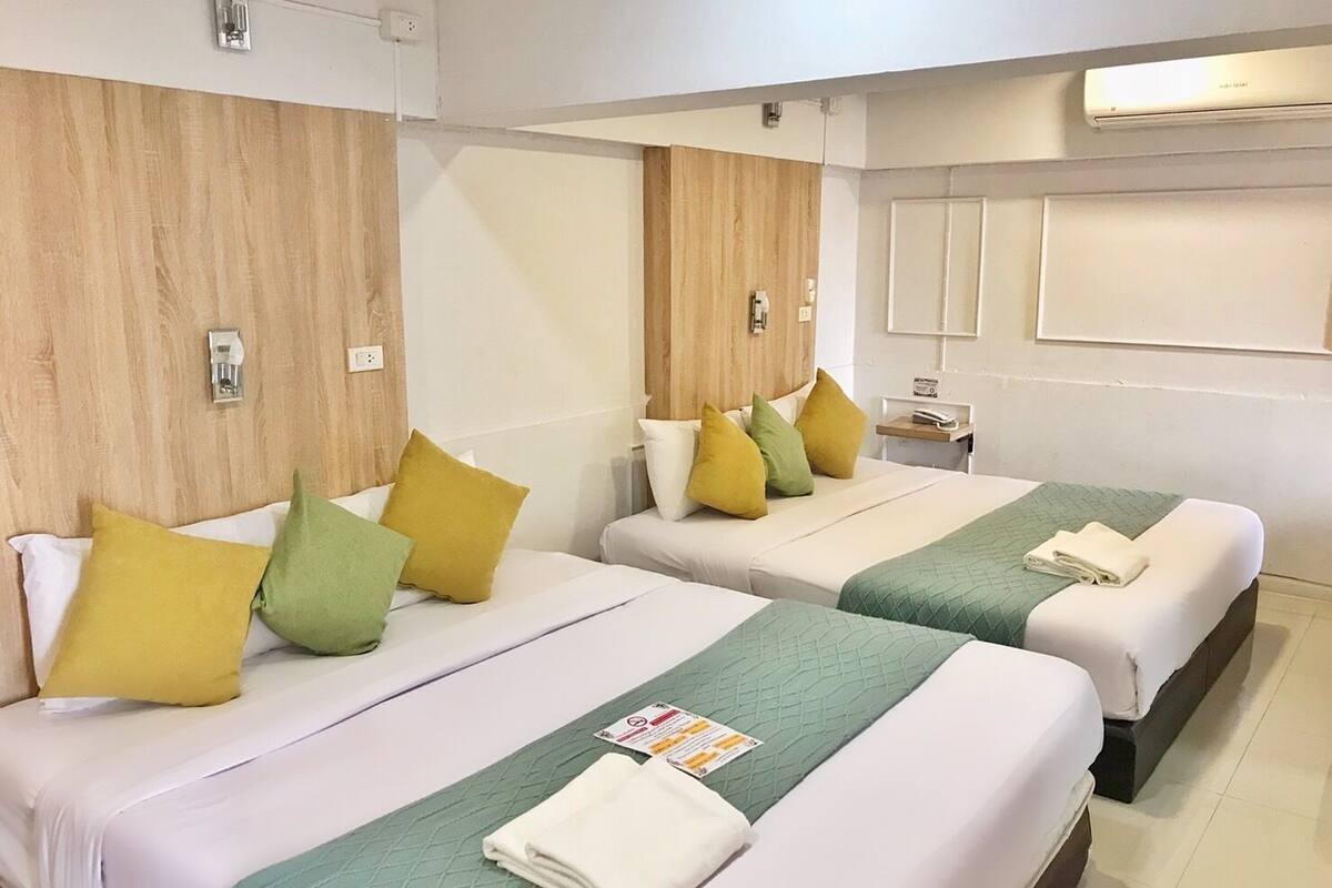 A hotel room with two beds, each with a green quilted bedspread and multiple pillows in yellow and green. There is a small bedside table with a lamp and a folded towel.