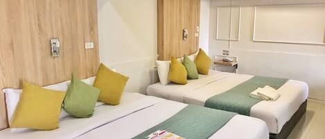 A hotel room with two beds, each with a green quilted bedspread and multiple pillows in yellow and green. There is a small bedside table with a lamp and a folded towel.