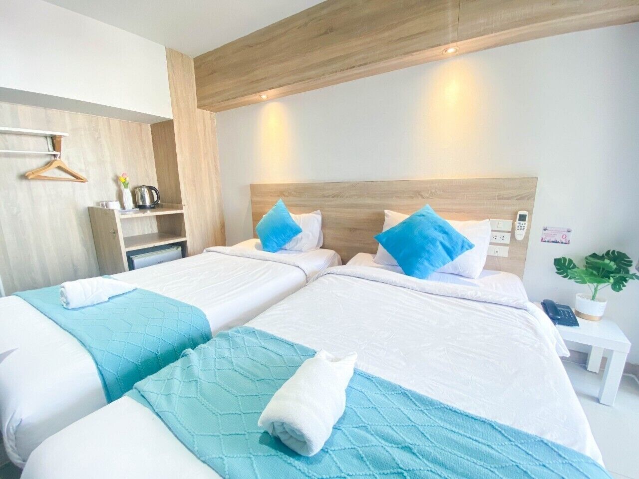 A hotel room with two beds, each with a blue quilted bedspread and white pillows. There is a wooden headboard, a bedside table with a phone, and a small plant on the nightstand.