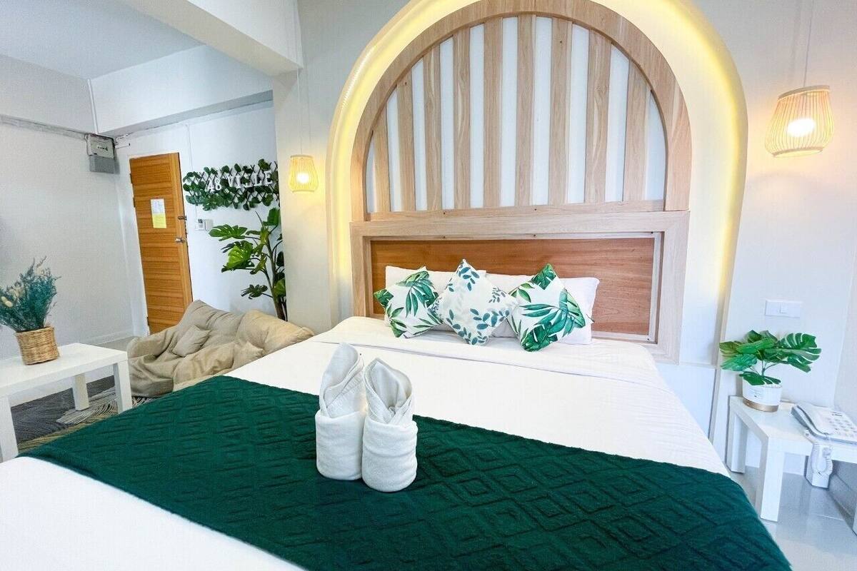 A hotel room with a bed, a desk, a chair, a plant, and a wall-mounted lamp.