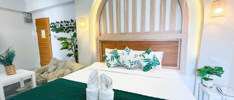 A hotel room with a bed, a desk, a chair, a plant, and a wall-mounted lamp.