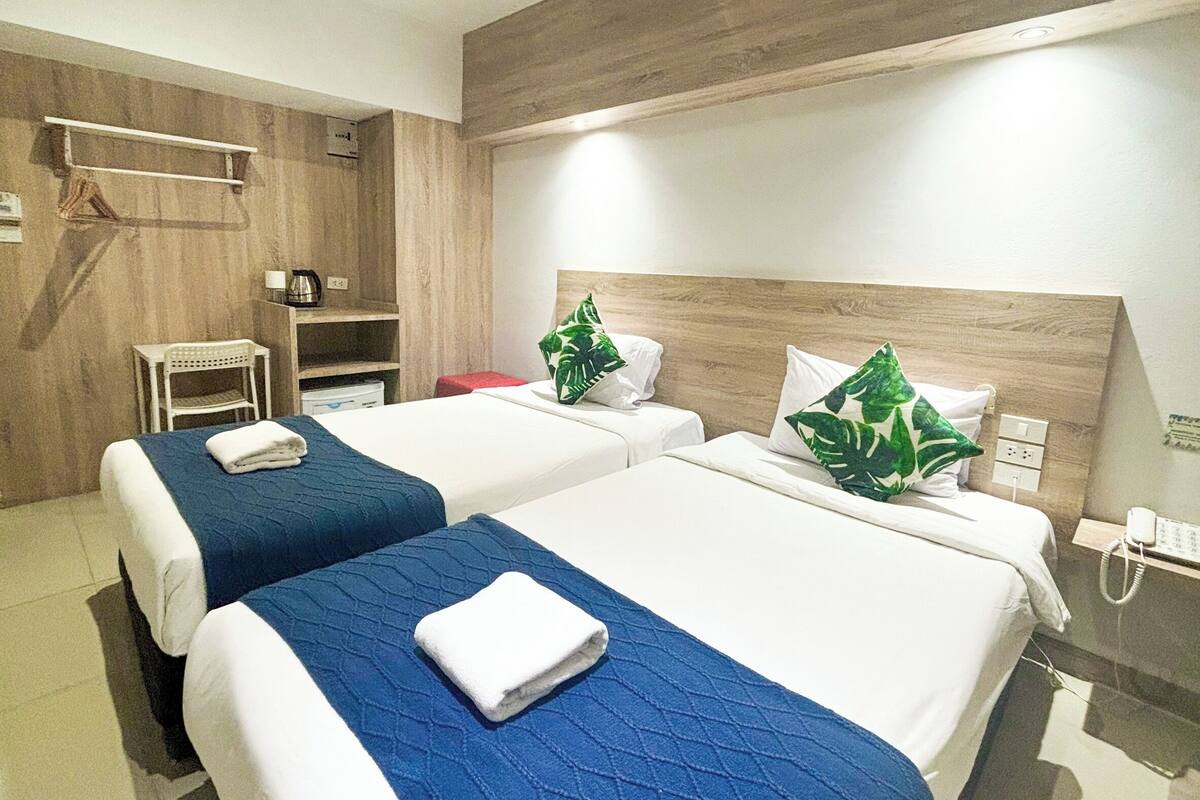 A hotel room with two beds, a wooden headboard, a small table, a chair, and a shelf with toiletries.