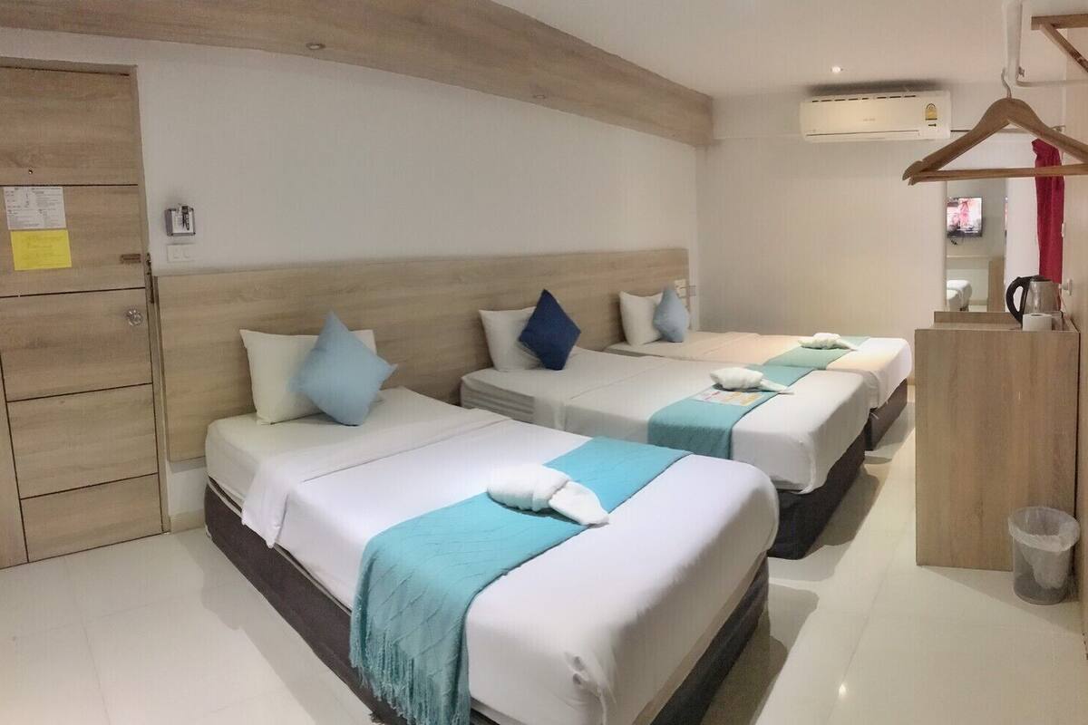 A hotel room with three beds, each with a white and blue bedspread, pillows, and a towel. There is a wooden wardrobe, a small trash bin, and an air conditioning unit on the wall.