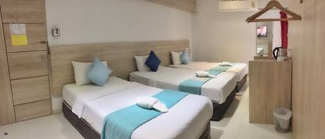 Une chambre d’hôtel avec trois lits, chacun recouvert d’une courtepointe blanche et bleue, avec des oreillers et une serviette. On y trouve une armoire en bois, une petite poubelle et un appareil de climatisation fixé au mur.