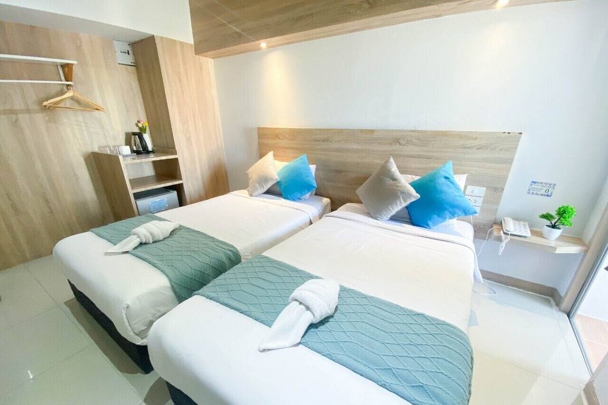 A modern hotel room with two beds, a wooden headboard, a bedside table with a plant, and a small shelf with a phone and a cup.