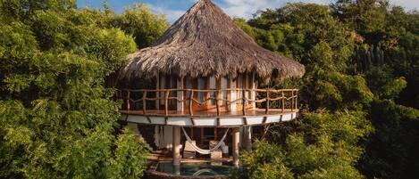 A treehouse with a thatched roof, wooden deck, and a pool.