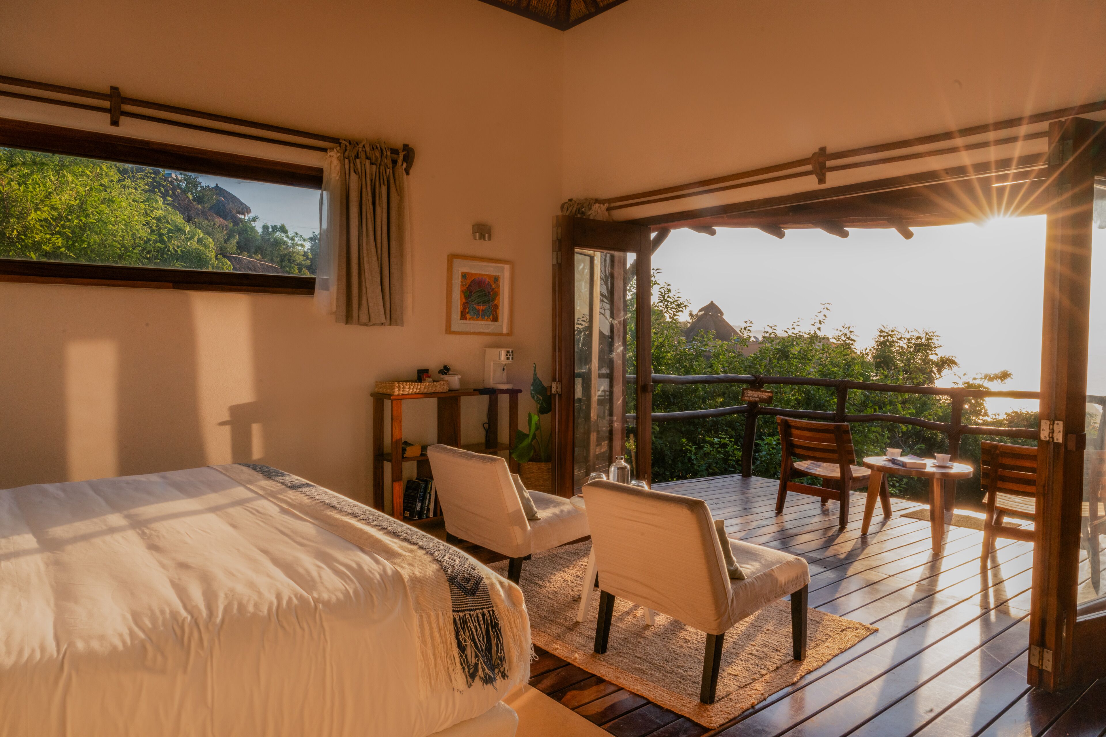 A bedroom with a large bed, a desk, and a chair, a balcony with wooden flooring, and a view of greenery and a mountain.