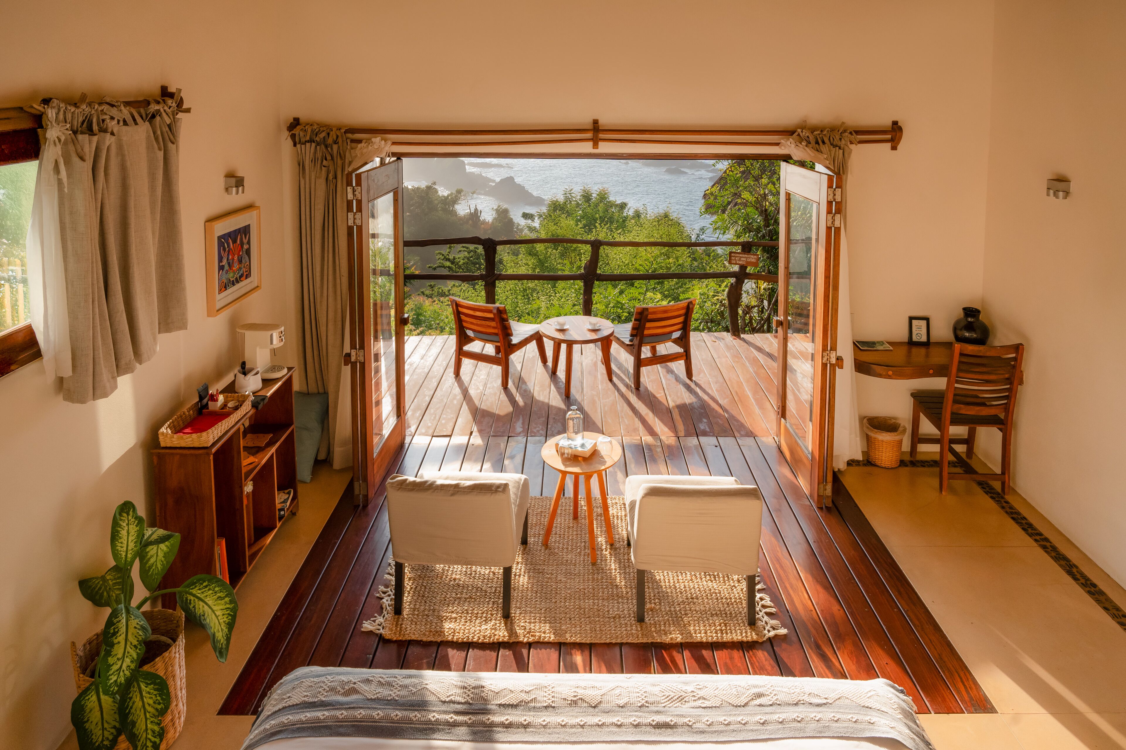 Una habitación con una cama, una mesita con sillas, un escritorio con una silla, una planta en maceta y vista al océano a través de un ventanal.