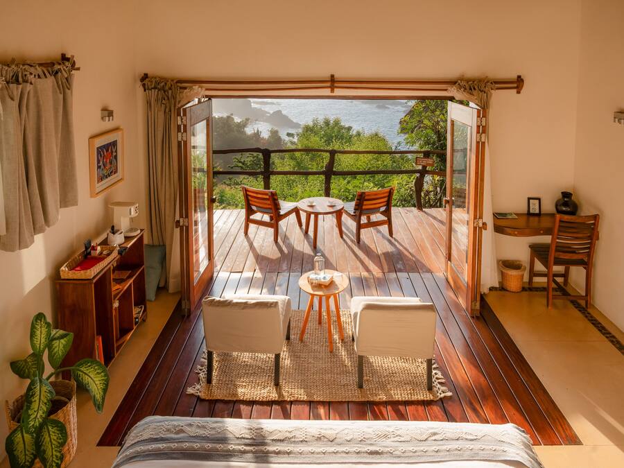 Una habitación con una cama, una mesita con sillas, un escritorio con una silla, una planta en maceta y vista al océano a través de un ventanal.