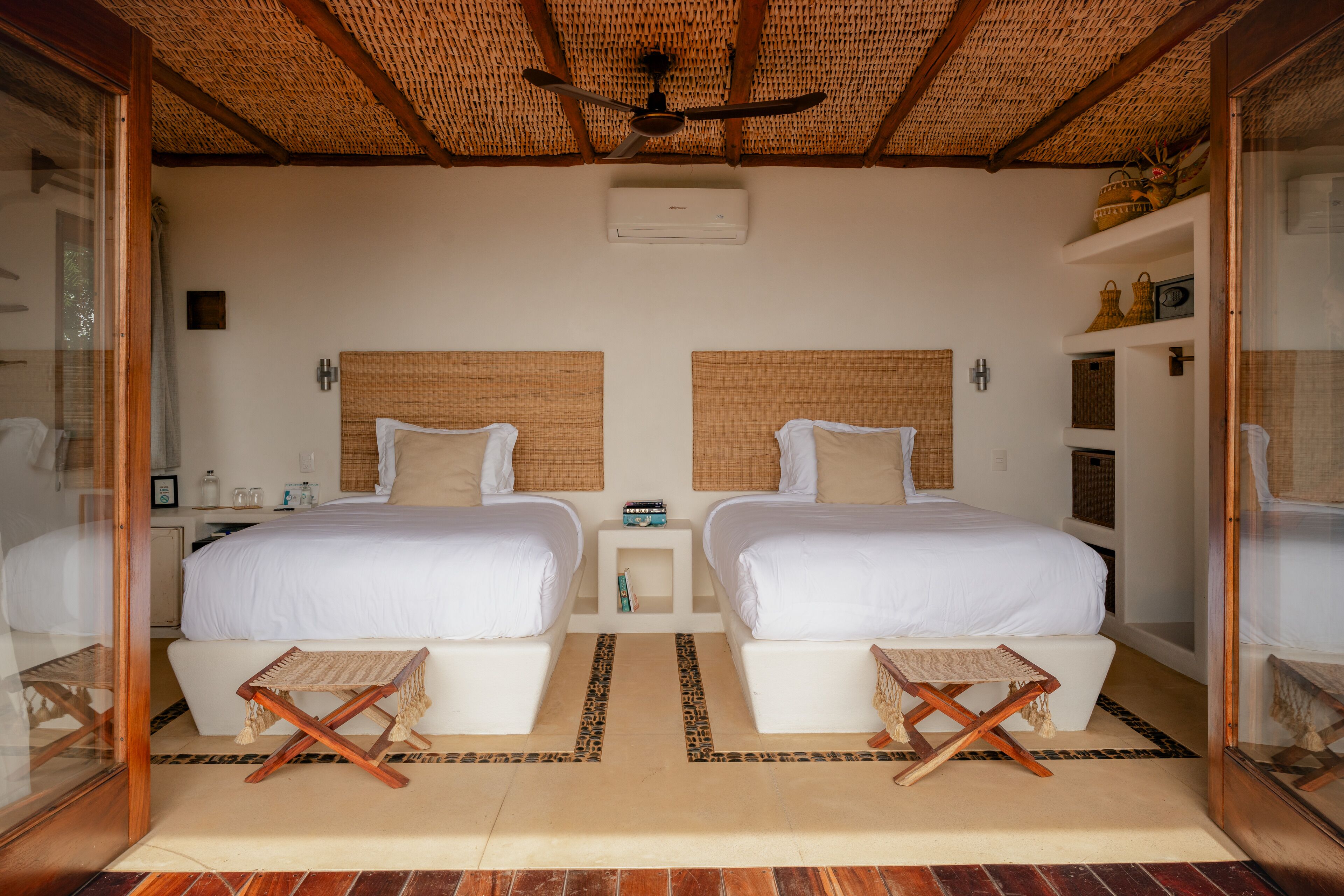 A room with two beds, a ceiling fan, a small table, and a shelf with decorative items.