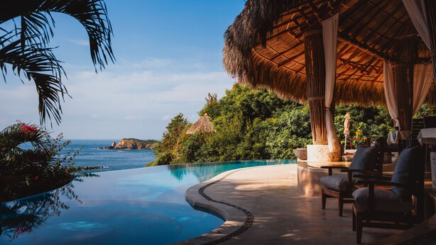 A poolside area with a curved infinity pool, a thatched-roof structure, and outdoor seating.