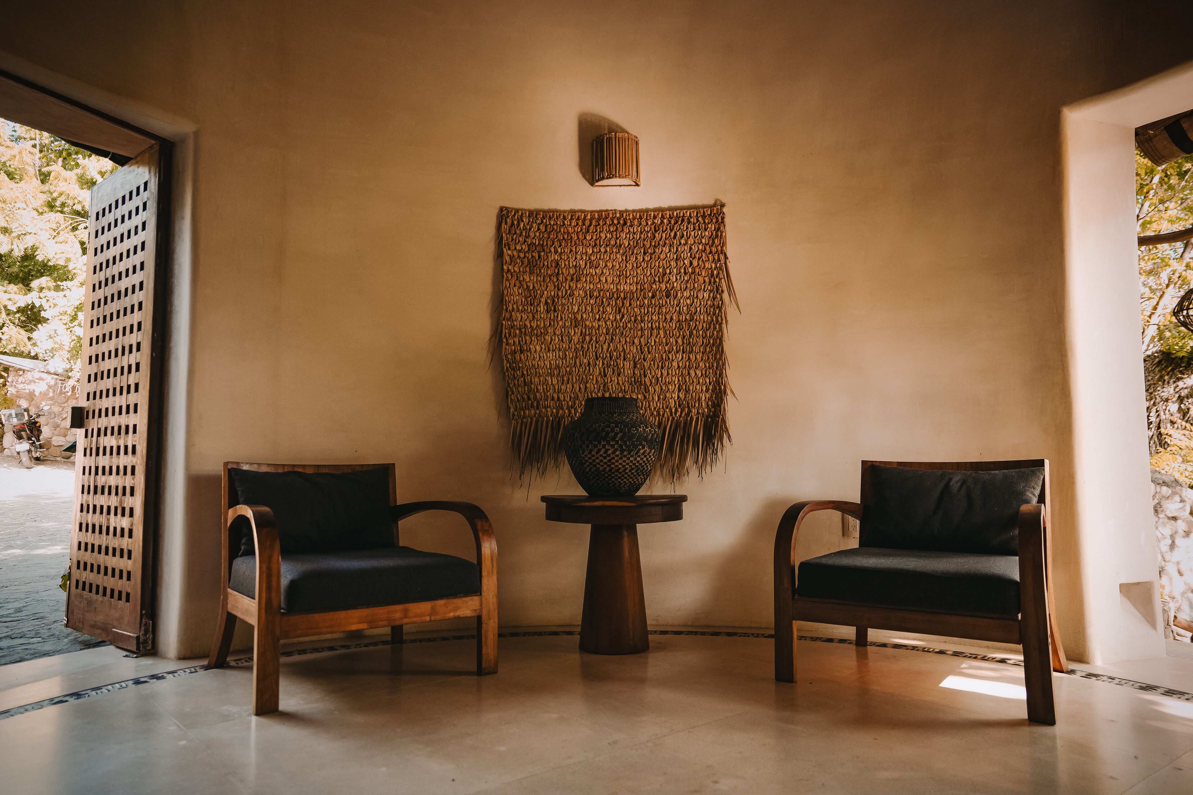 Um quarto com duas cadeiras de madeira, um painel de parede tecido e uma pequena mesa com um vaso.