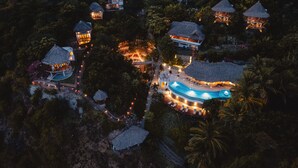 An aerial view of a resort with multiple thatched-roof villas, a swimming pool, and a well-lit pathway.
